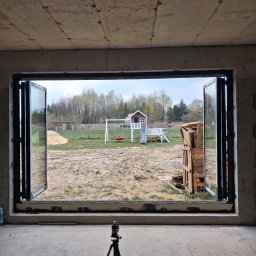 P&Home - Rozłożone, czarne drzwi harmonijkowe z widokiem na plac zabaw i ogród, zamontowane w surowym, betonowym otworze. Na pierwszym planie niwelator laserowy na statywie.