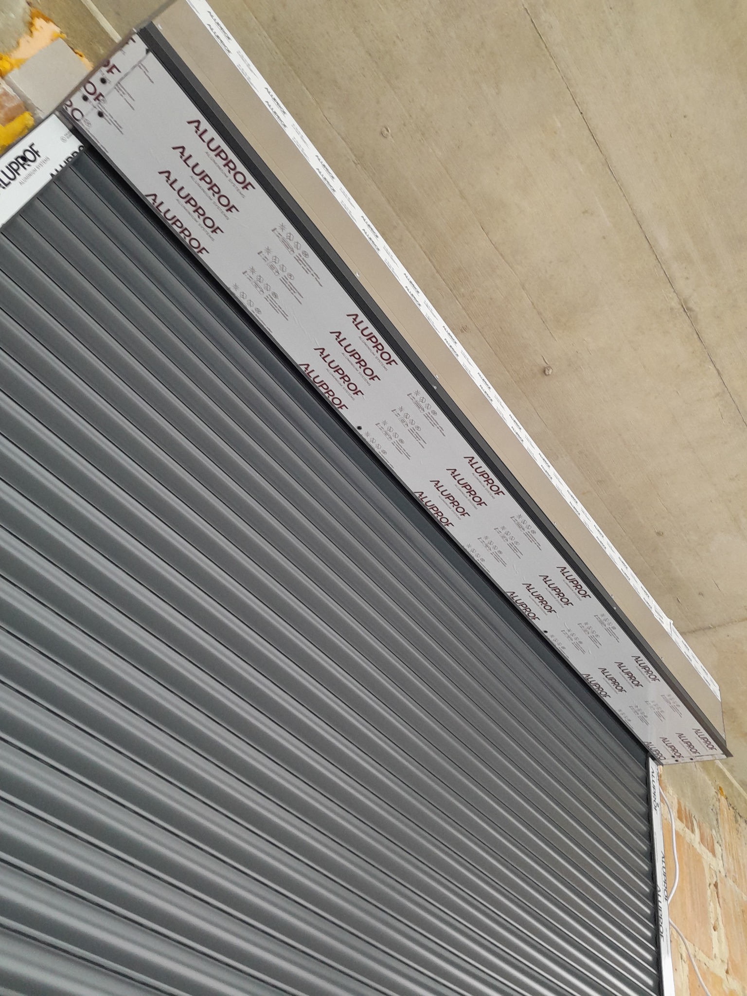 Szara roleta zewnętrzna Alprof, częściowo zwinięta, zamontowana na tle surowego muru. Widoczne detale konstrukcyjne i logo producenta.