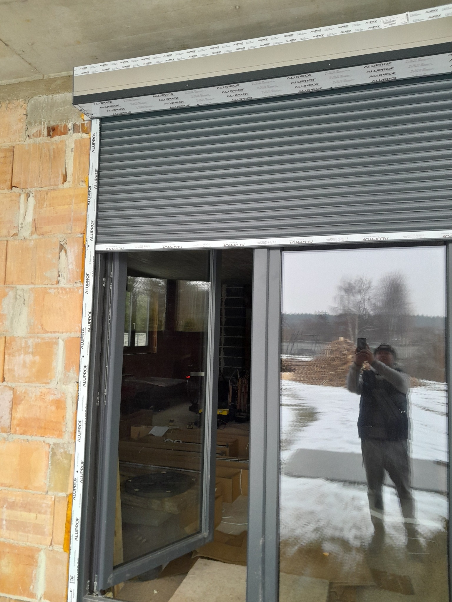 Szara roleta zewnętrzna Aluplast zamontowana na oknie z widocznym odbiciem osoby robiącej zdjęcie. Surowy mur z cegły i betonowy strop w tle.