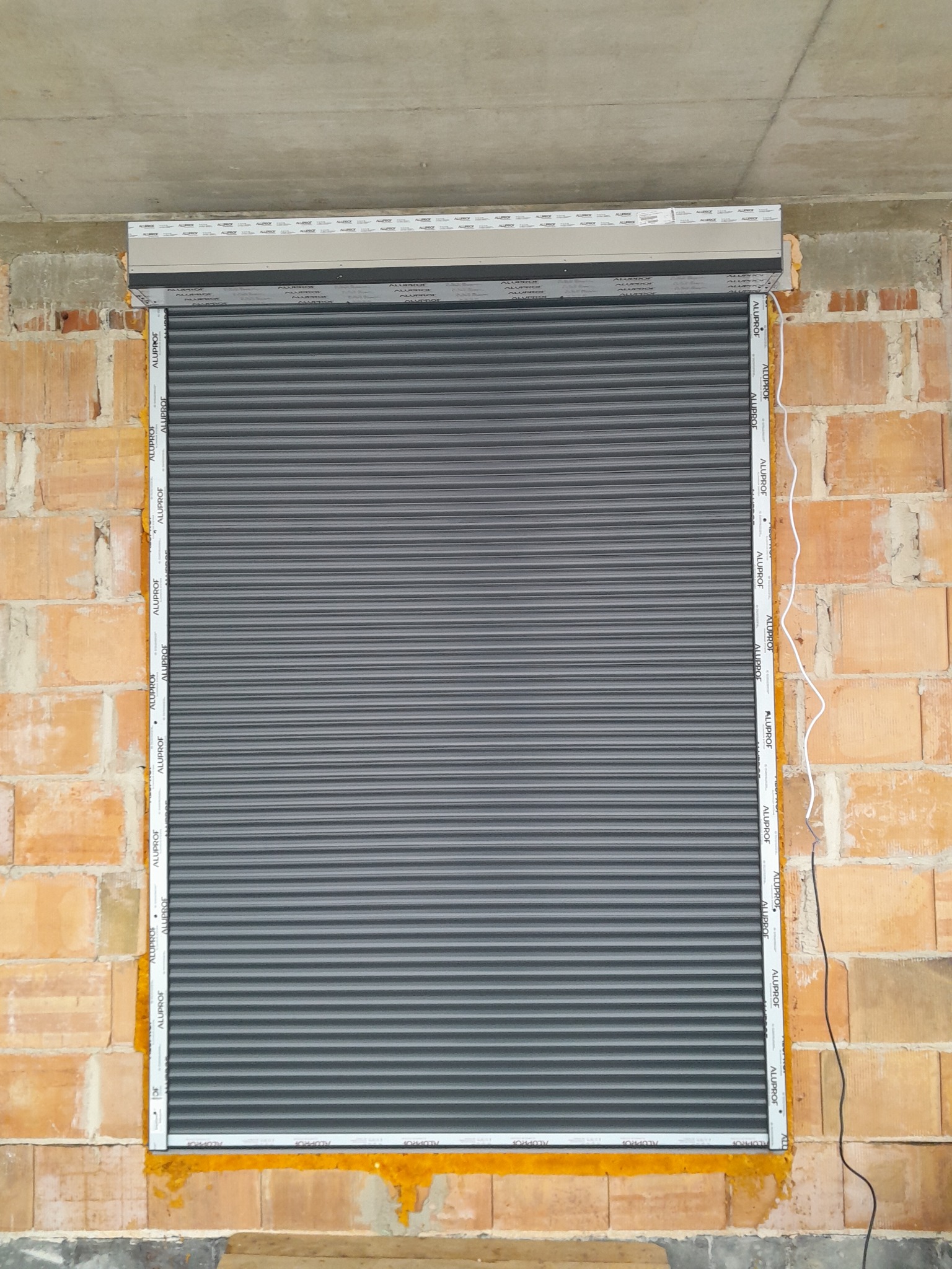 Szara roleta zewnętrzna Aluplrof, zamontowana w surowym otworze okiennym z cegły, widoczne przewody elektryczne. Detal montażu.