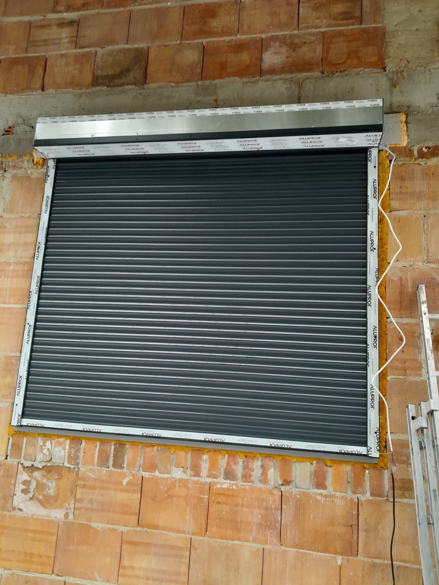 Szara roleta zewnętrzna zamontowana w oknie budynku z cegły, widoczne elementy montażowe i pianka uszczelniająca. Rama okienna z napisem 'ALUPROF'.