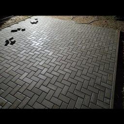 Rzk Kamyk - Ułożona, szara kostka brukowa w jodełkę, z kilkoma kostkami obok, gotowa do ułożenia. Widoczne obrzeża i fragment trawnika. Nocne oświetlenie.