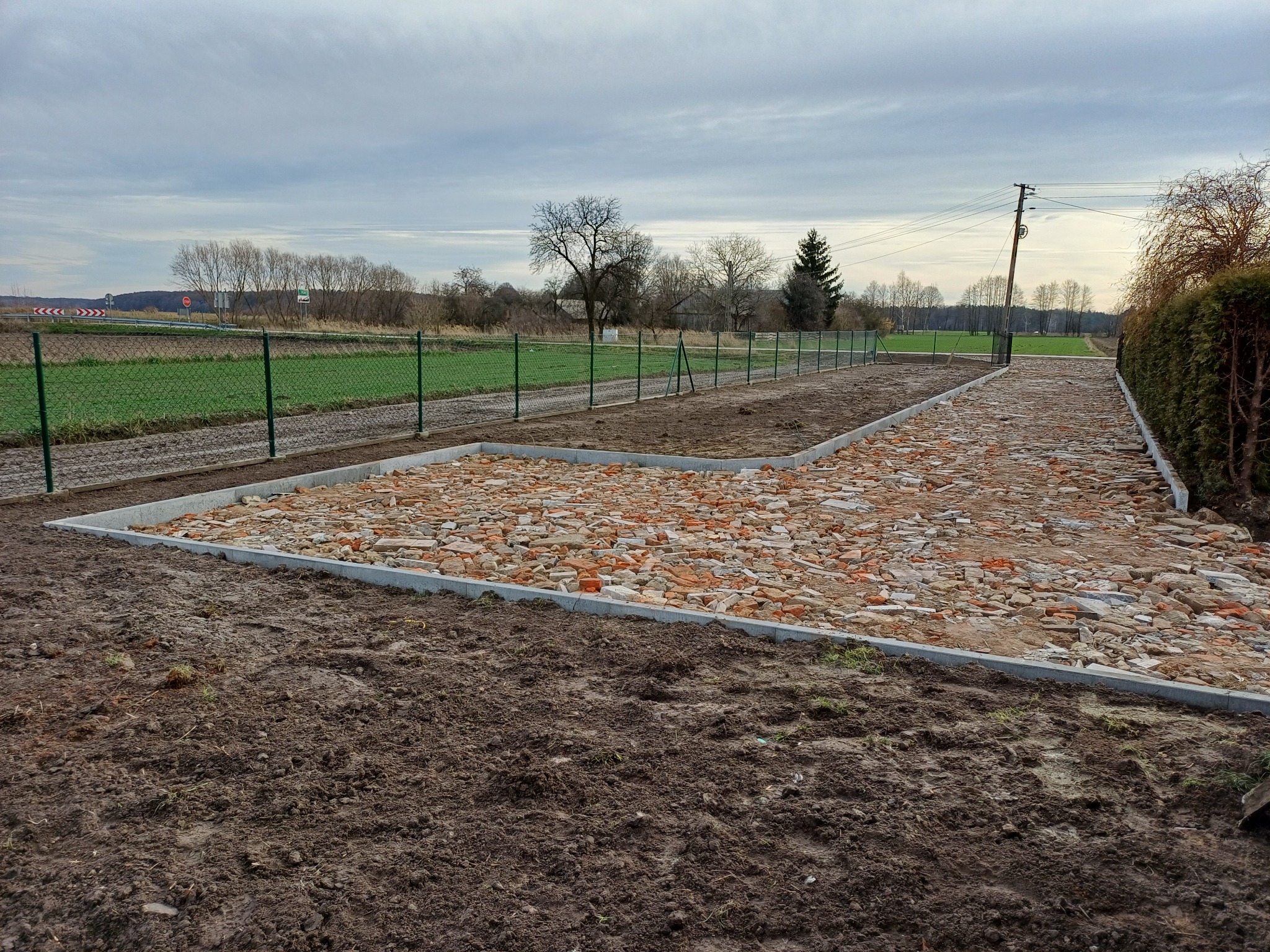 Przygotowany teren pod budowę z obrzeżami i gruzem ceglanym, widok na zielone pola i drzewa w tle, pochmurne niebo. Obrzeża betonowe wyznaczają kształt przyszłej konstrukcji.