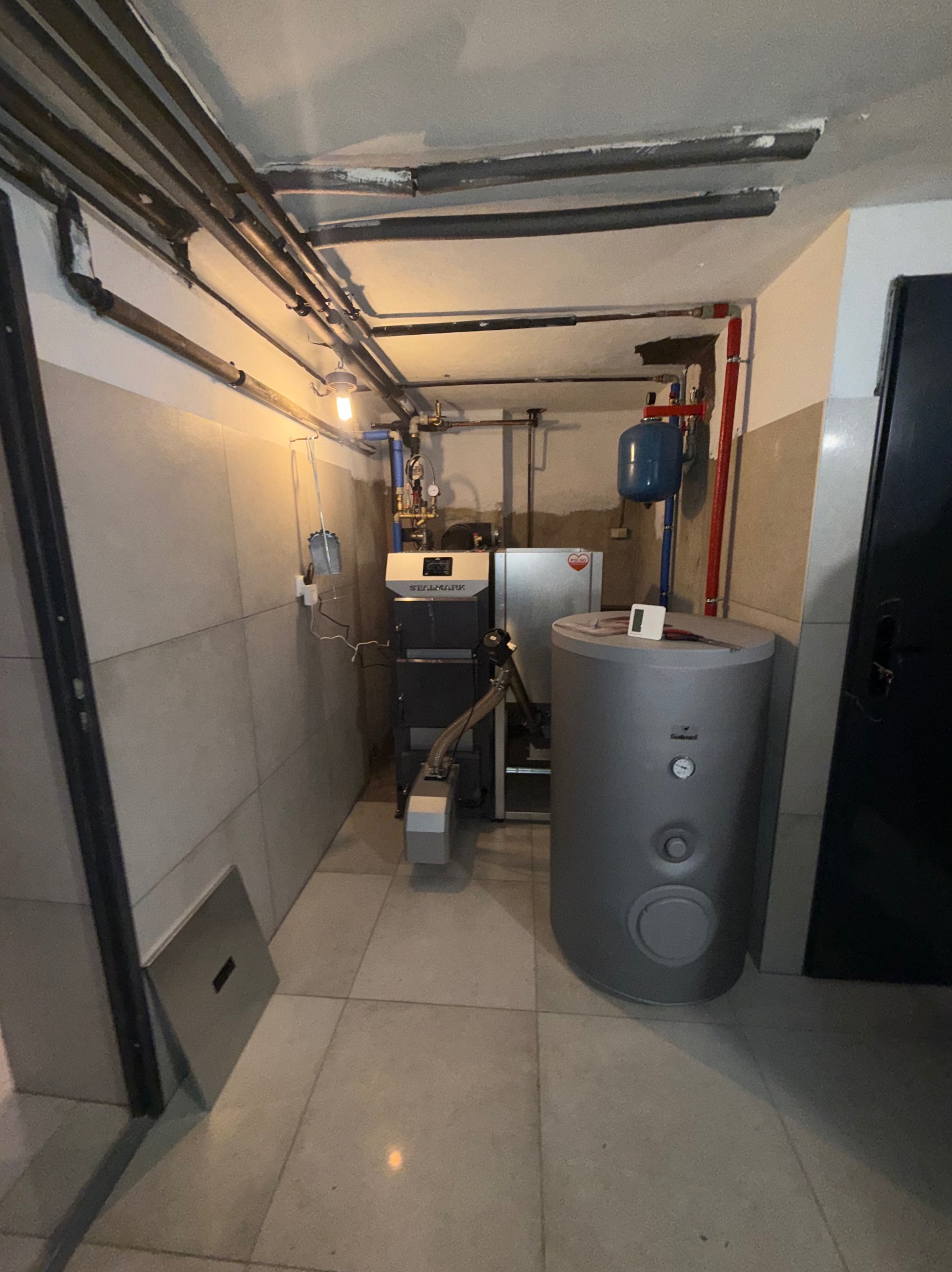 Kotłownia z piecem na paliwo stałe marki SEALMARK i szarym zasobnikiem ciepłej wody. Widoczne rury instalacji grzewczej pod sufitem i płytki na ścianach.