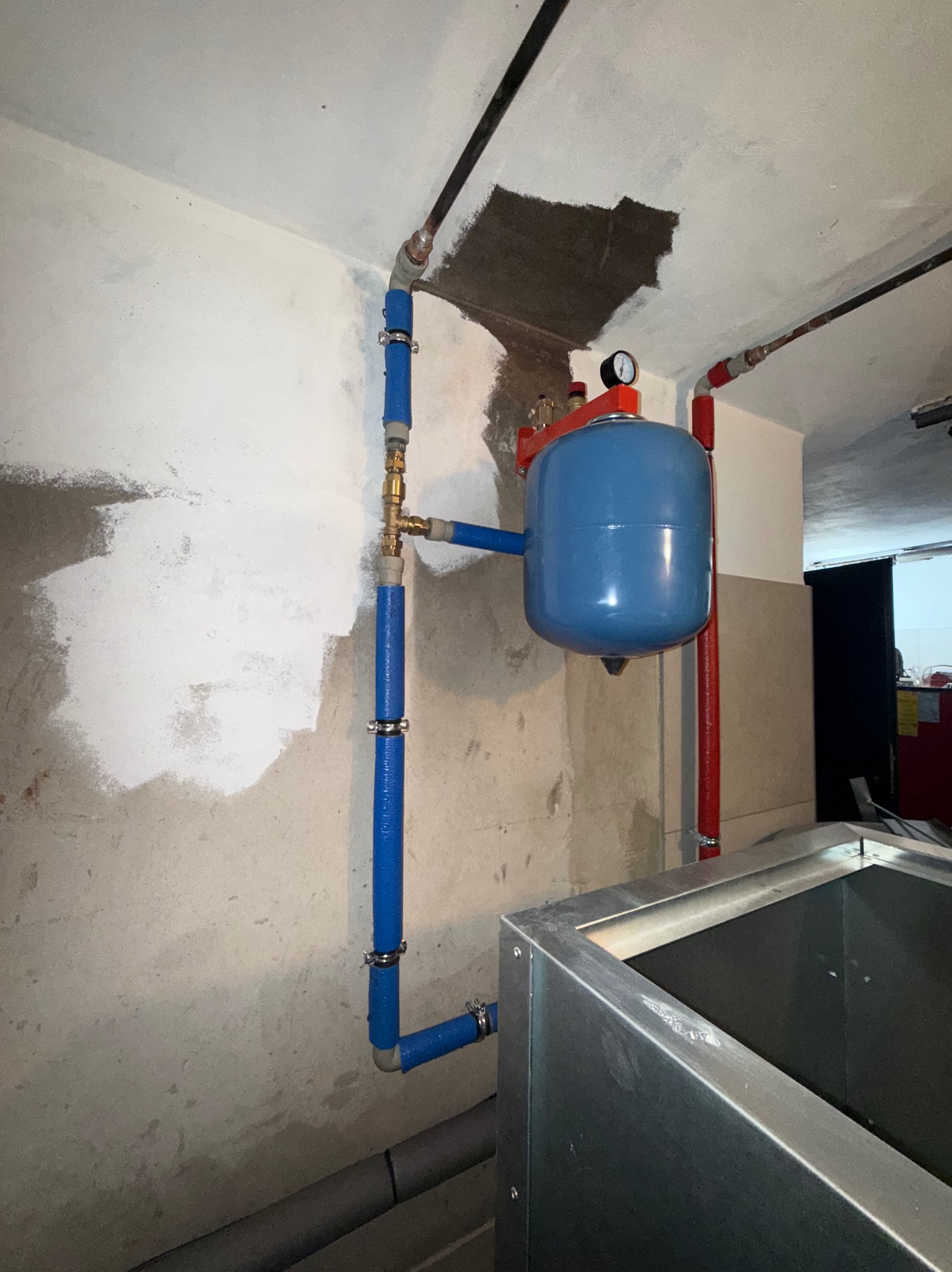 Niebieski zbiornik ciśnieniowy z manometrem, zamontowany na ścianie z instalacją z rur w kolorze niebieskim i czerwonym. Widoczne ślady po naprawach na ścianie.