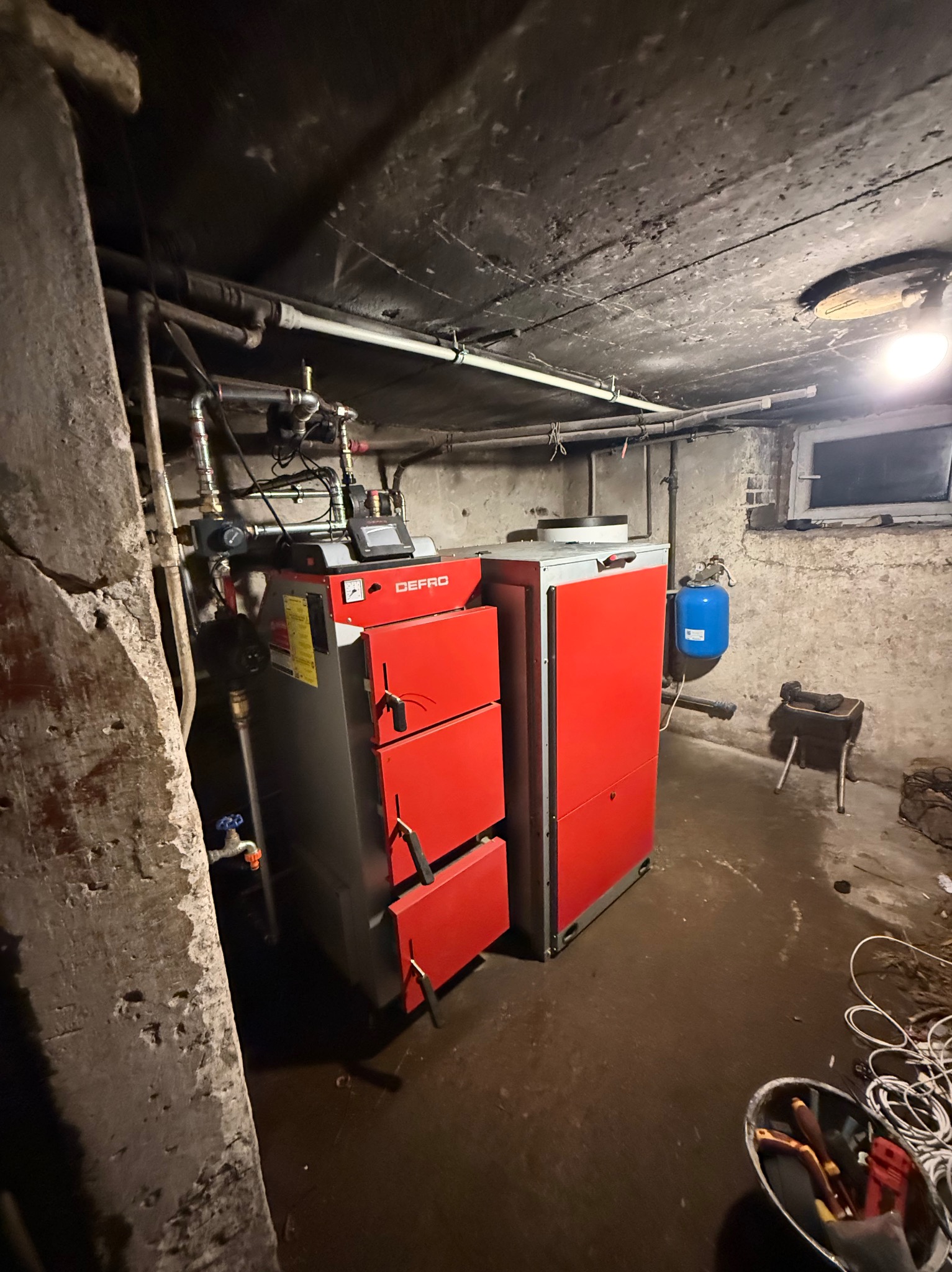 Czerwono-szary piec DEFRO na paliwo stałe w piwnicy, widoczne rury instalacji, zbiornik wyrównawczy i narzędzia. Surowy betonowy strop i ściany.