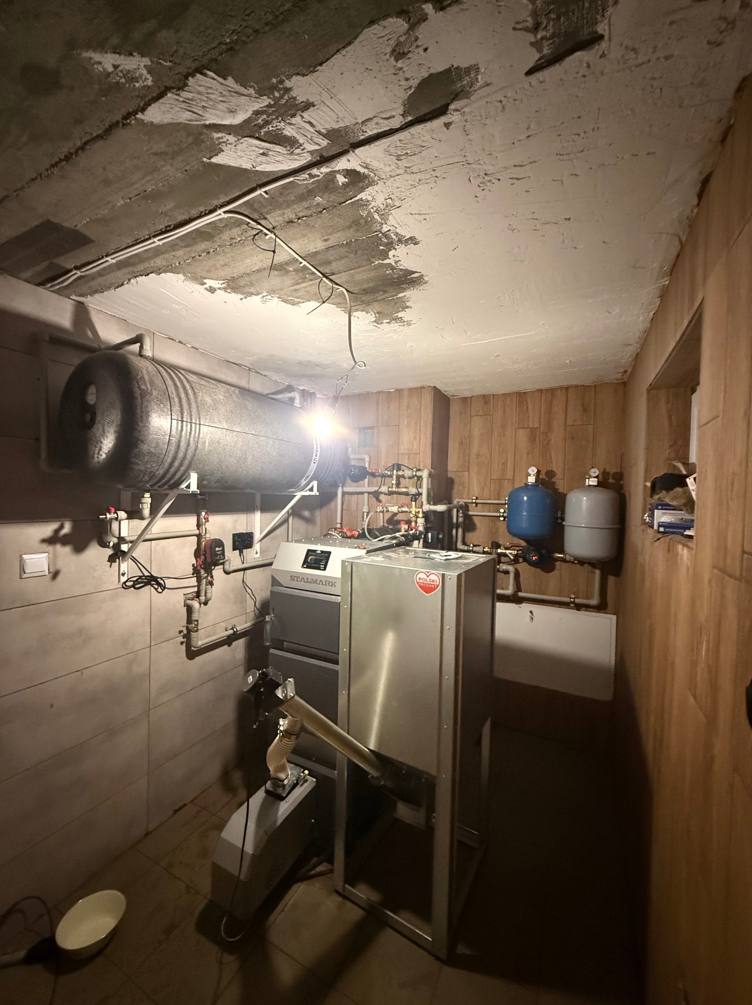Kotłownia z piecem Stalmark, zbiornikiem buforowym i instalacją hydrauliczną. Widoczne rury, zawory i osprzęt. Ściany wyłożone płytkami i boazerią.