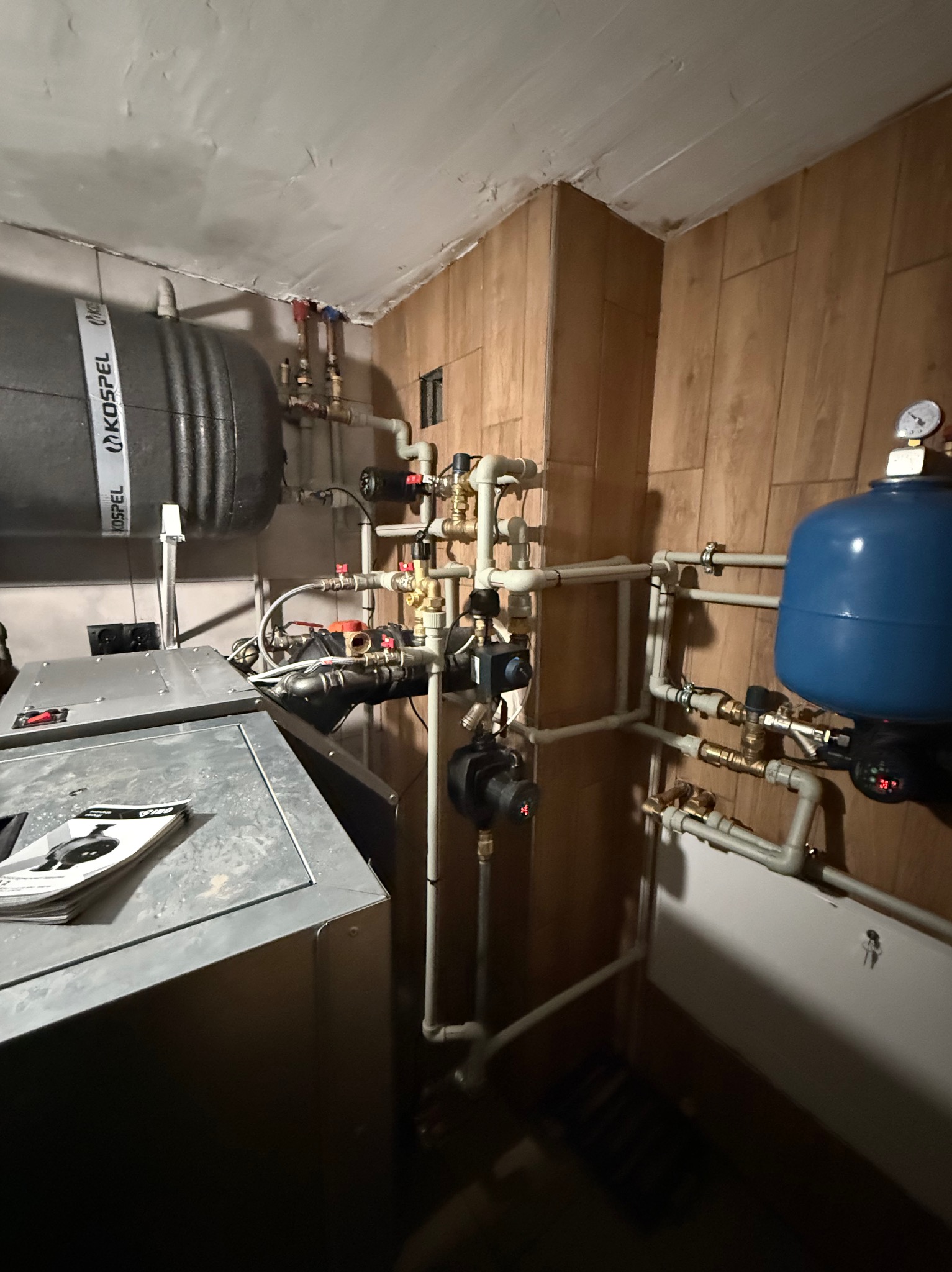 Kotłownia z piecem Kospel, zbiornikiem buforowym i instalacją hydrauliczną z licznymi zaworami, rurami z tworzywa i mosiężnymi złączkami w pomieszczeniu z boazerią.