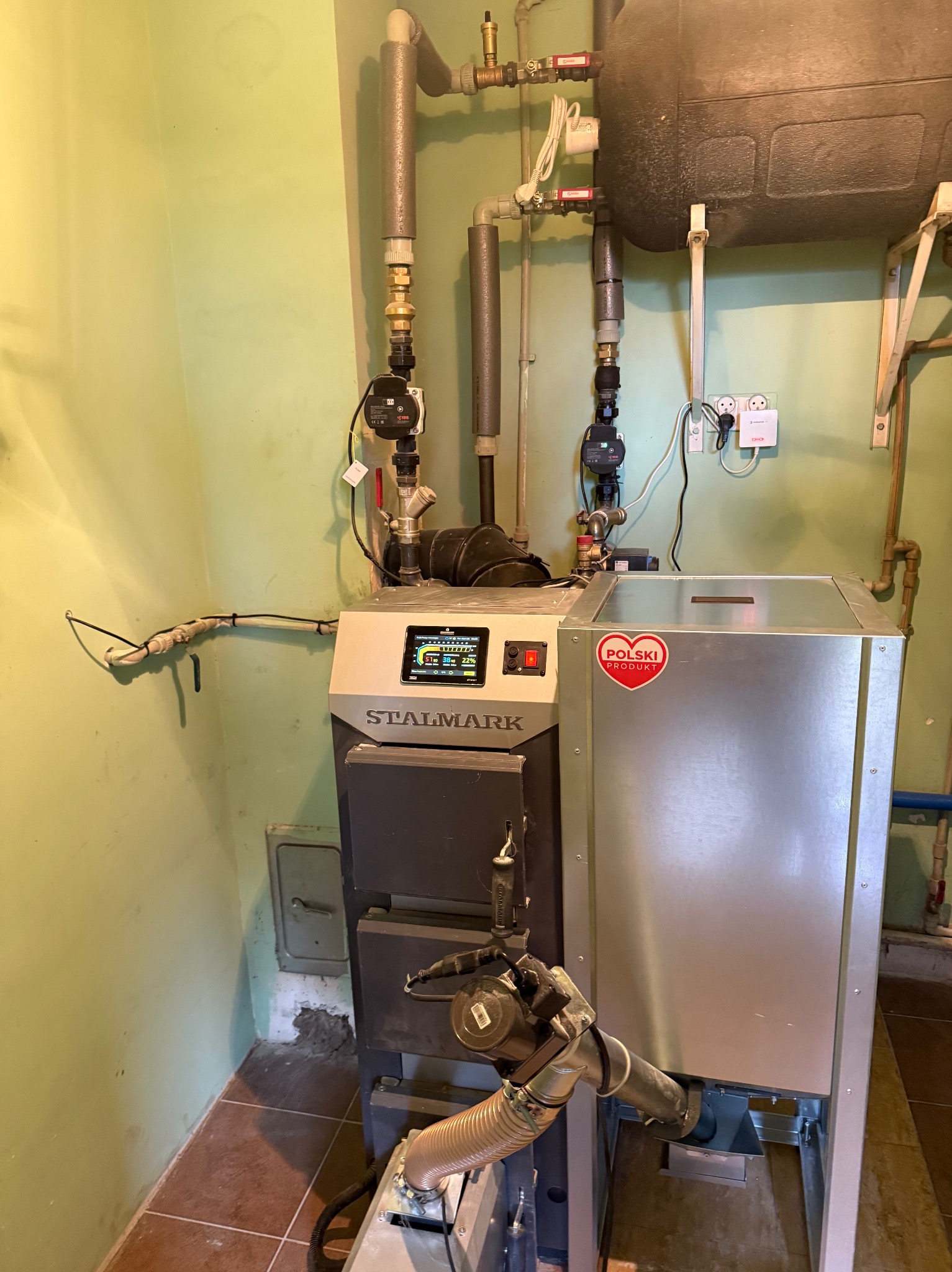 Zamontowany piec na paliwo stałe marki Stalmark z widocznymi rurami, pompami i zbiornikiem wyrównawczym w kotłowni o zielonych ścianach i brązowej podłodze.