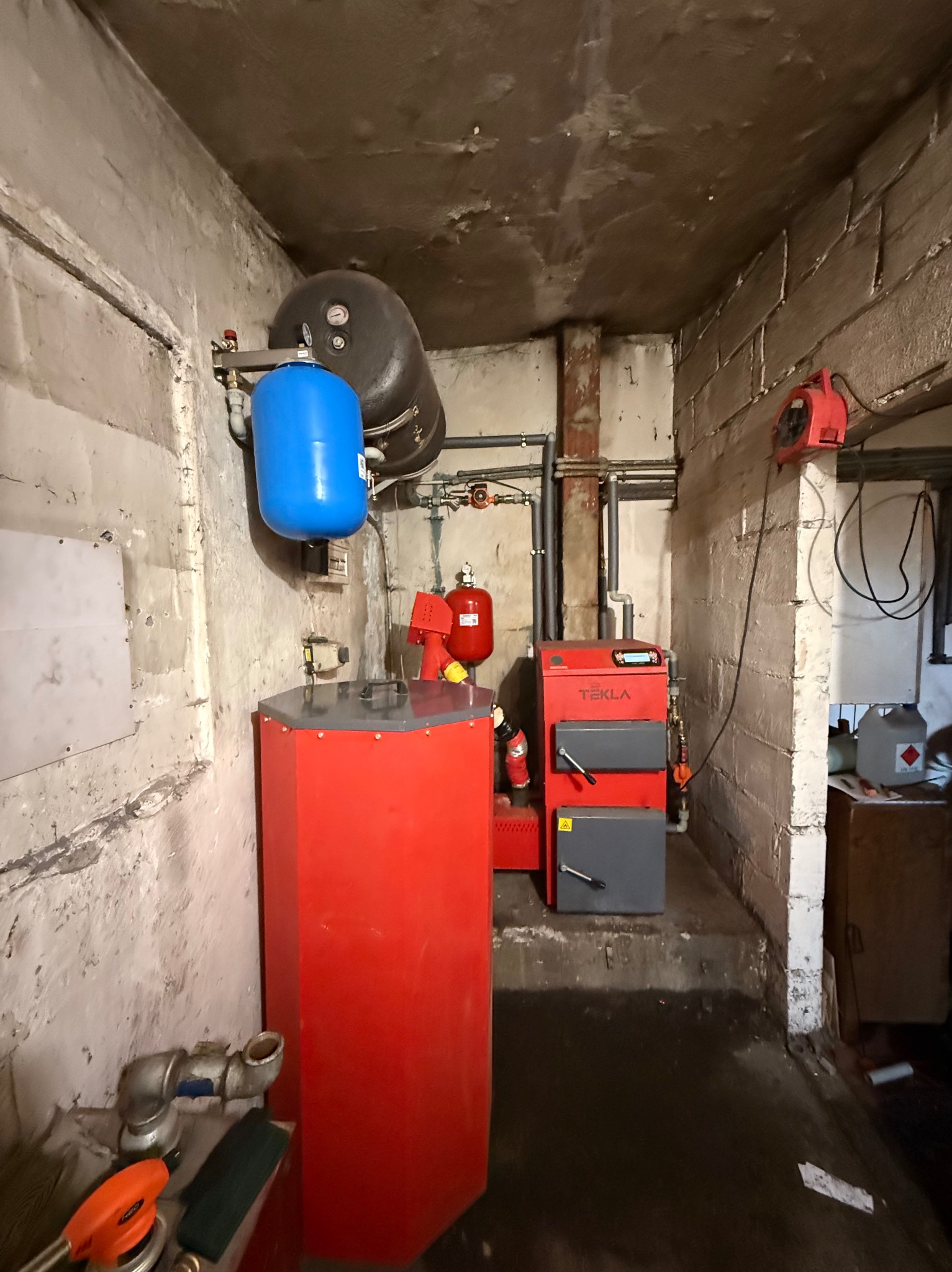 Czerwony piec Tekla w kotłowni z szarymi drzwiczkami, zbiornik wyrównawczy i instalacja. Widoczne rury i elementy hydrauliczne. Ściany z surowego betonu.