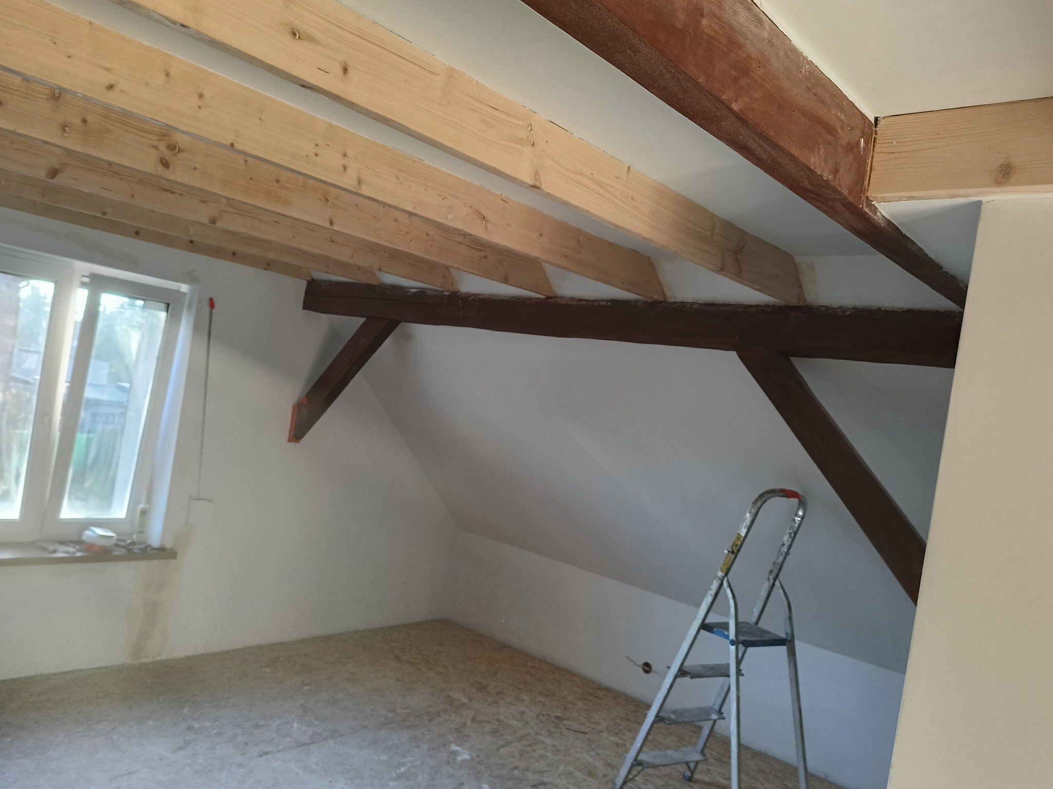 Adaptacja poddasza: surowe drewniane belki stropowe, białe ściany, okno i drabina. Widoczny etap prac wykończeniowych, surowy stan podłogi z płyty OSB.