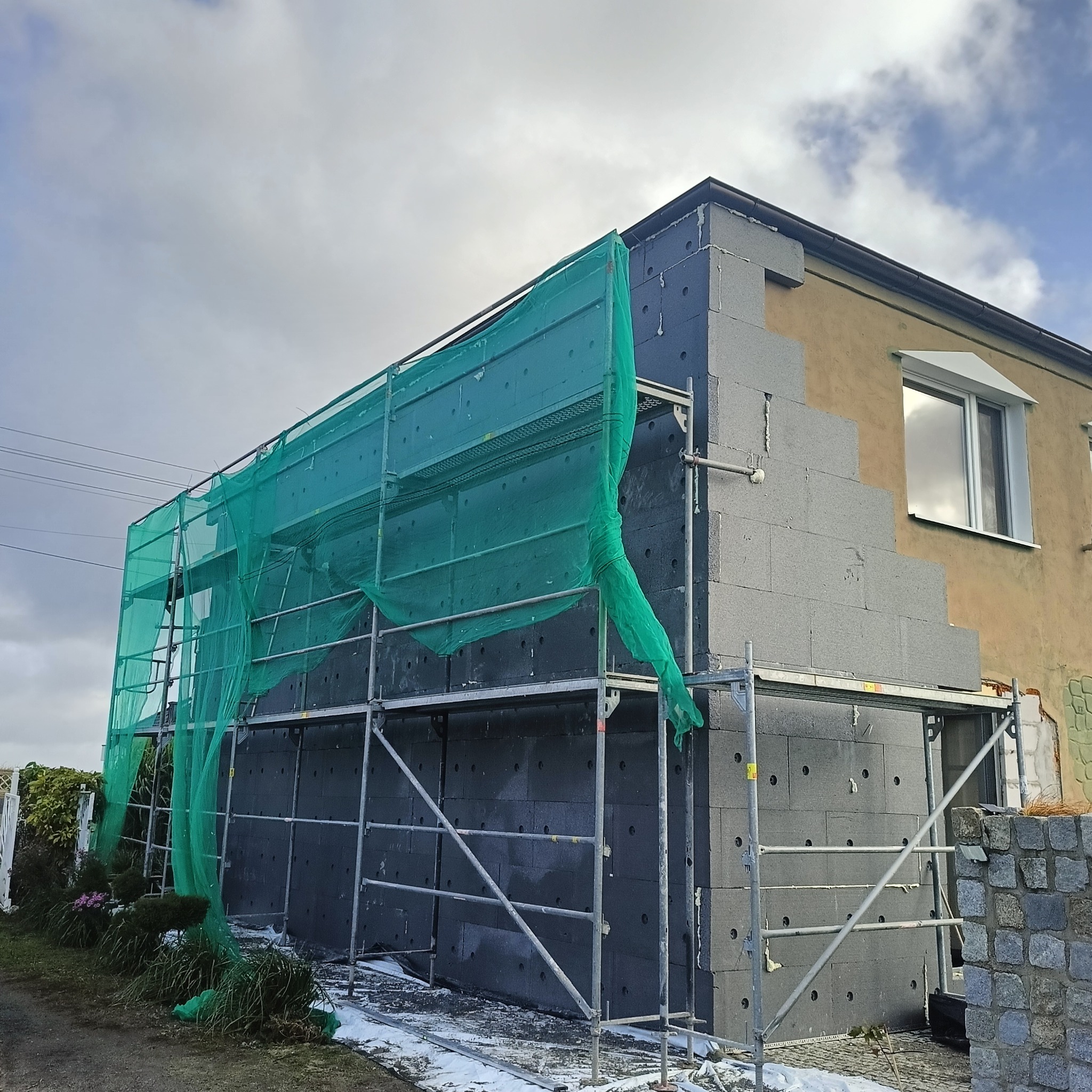 Ocieplanie elewacji domu z użyciem grafitowego styropianu, widoczne rusztowanie i siatka ochronna. Częściowo ocieplona ściana kontrastuje z fragmentem starej elewacji.