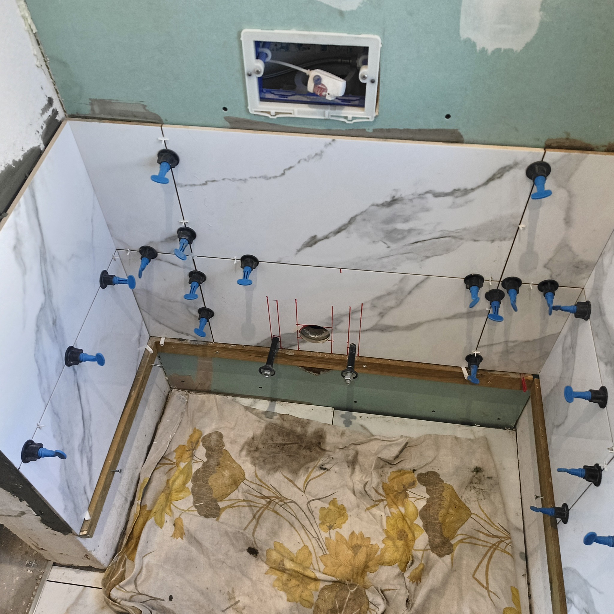 Montaż płytek imitujących marmur w łazience, z użyciem systemu poziomowania. Widoczna instalacja podtynkowa i zabezpieczona podłoga. Ujęcie z góry.