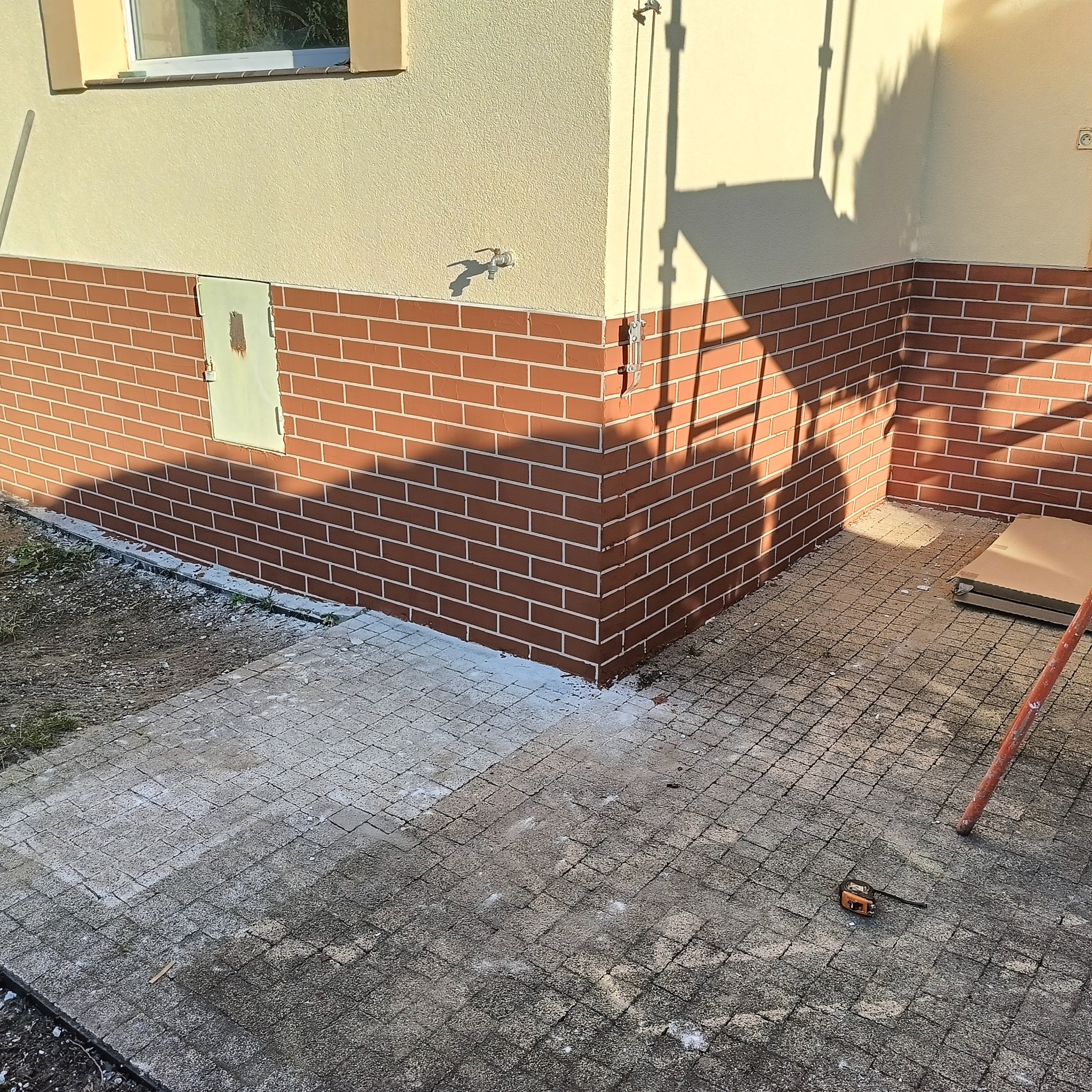 Elewacja budynku z ceglaną okładziną i jasnym tynkiem, widoczny fragment chodnika z kostki, cień rusztowania. Detal wykończenia zewnętrznego.