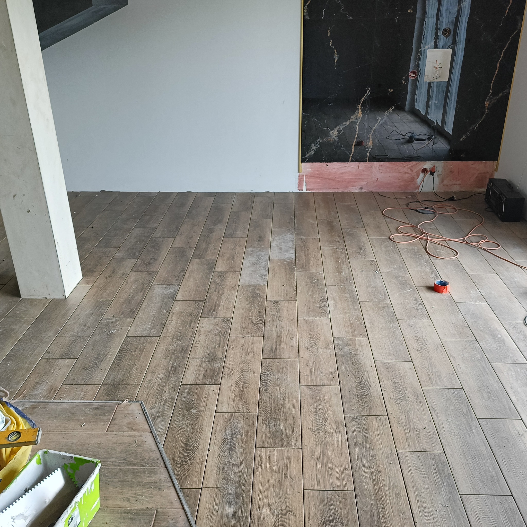 Układanie płytek podłogowych imitujących drewno w jasnym odcieniu. W tle ściana z czarnym marmurem i widoczne narzędzia pracy.