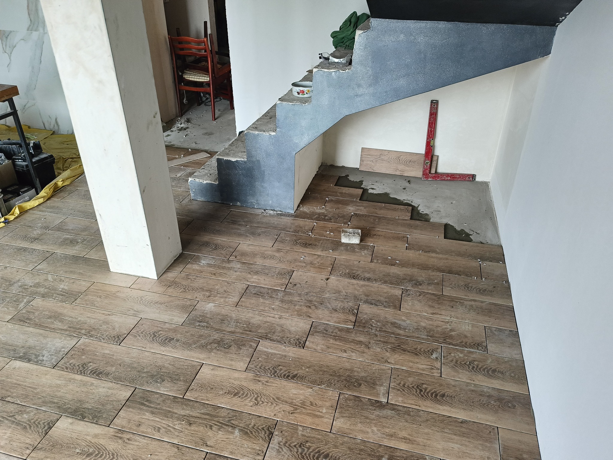 Układanie drewnopodobnych płytek na podłodze przy schodach. Widoczny fragment kolumny, poziomica i płytki ułożone w jodełkę. Prace wykończeniowe w trakcie realizacji.