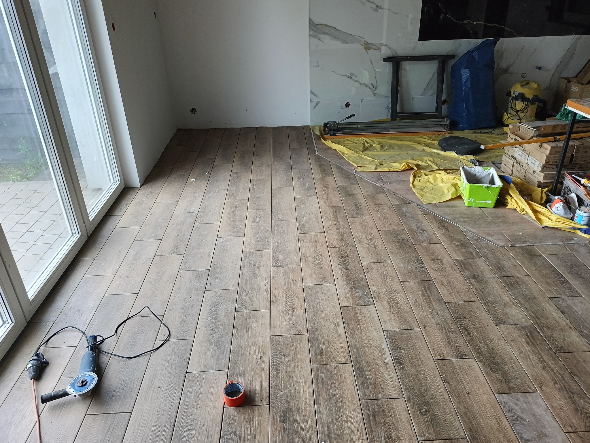 Ułożona podłoga z płytek imitujących drewno w jasnym pomieszczeniu z dużym oknem. Widoczne narzędzia pracy i zabezpieczenia podłoża folią.