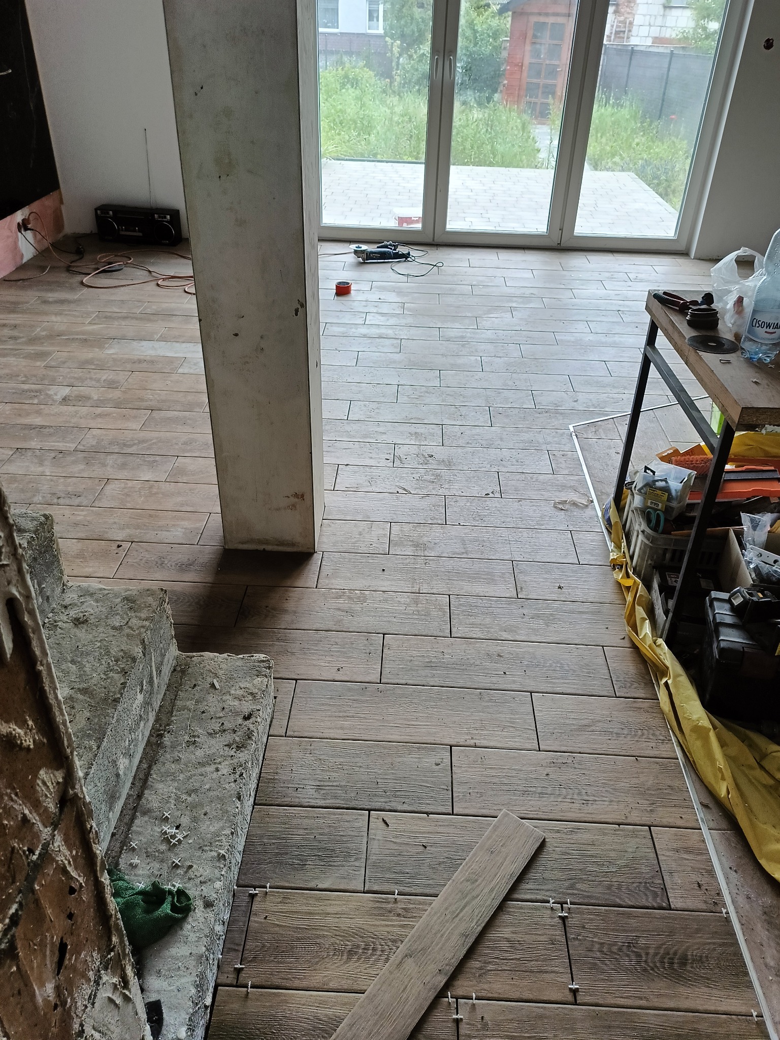 Układanie podłogi z płytek imitujących drewno w jasnym wnętrzu z dużymi oknami. Widoczne narzędzia i materiały budowlane. Etap prac wykończeniowych.