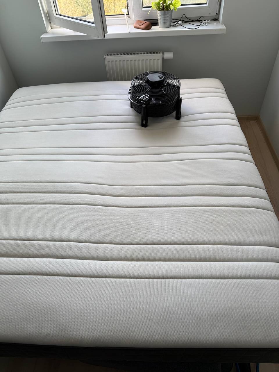 Materac z wentylatorem do suszenia po praniu, ustawiony na łóżku w pokoju z oknem i grzejnikiem. Widok z góry, neutralne oświetlenie.