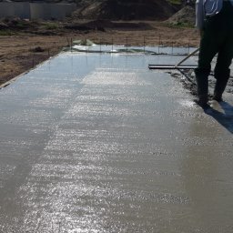 URB usługi remontowo-budowlane - Wyrównywanie mokrej wylewki betonowej przez pracownika w zielonym kombinezonie. W tle widoczne szalunki i hałda ziemi.