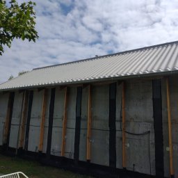 URB usługi remontowo-budowlane - Ściana budynku w trakcie budowy z widocznymi elementami konstrukcyjnymi i izolacją, blaszany dach, fragment zieleni i niebo z chmurami w tle.