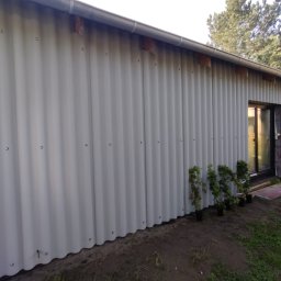 URB usługi remontowo-budowlane - Ściana budynku obłożona blachą falistą, widoczne mocowania. Przy ścianie ustawione doniczki z roślinami. W tle drzewa.