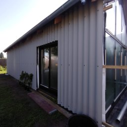 URB usługi remontowo-budowlane - Budowa domu szkieletowego z szarą blachą falistą na elewacji, widoczne okno i folia izolacyjna Tyvek. Ujęcie z zewnątrz, w tle zieleń i fragment dachu.