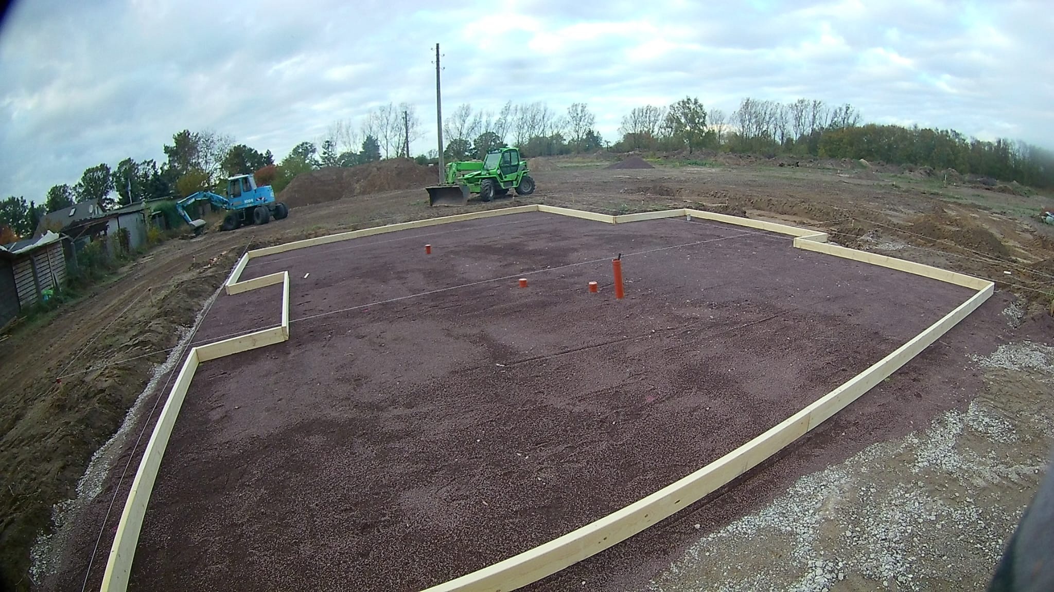 Przygotowany teren pod fundamenty z drewnianym szalunkiem, widoczne maszyny budowlane w tle. Grunt wyrównany, gotowy do dalszych prac.