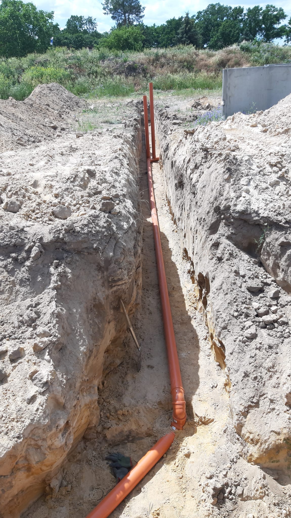 Pomarańczowe rury kanalizacyjne ułożone w wykopie ziemnym, widok z góry. W tle zielona roślinność i betonowy element konstrukcyjny.