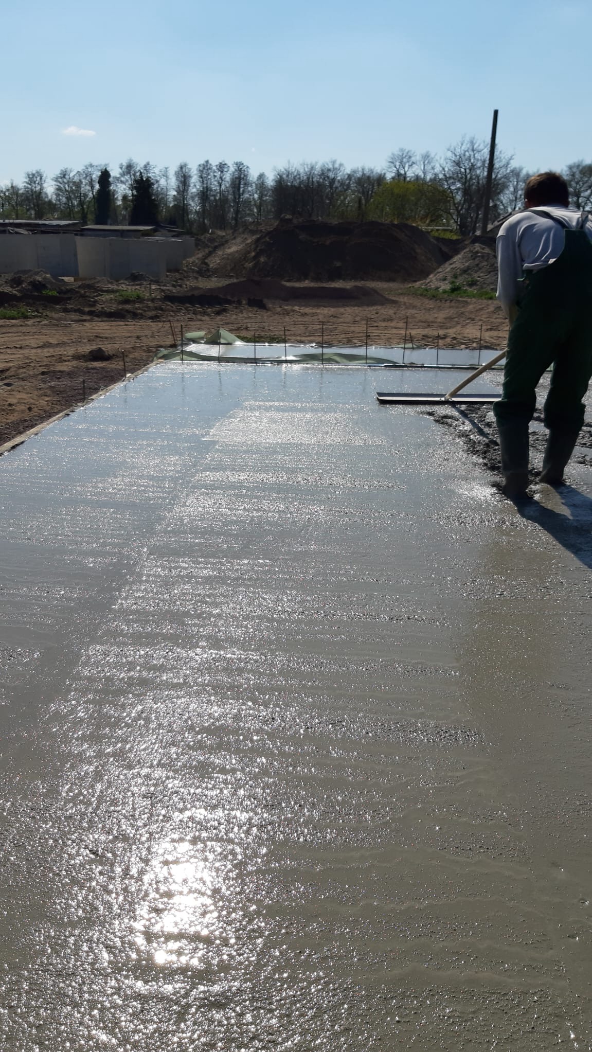 Wyrównywanie mokrej wylewki betonowej przez pracownika w zielonym kombinezonie. W tle widoczne szalunki i hałda ziemi.