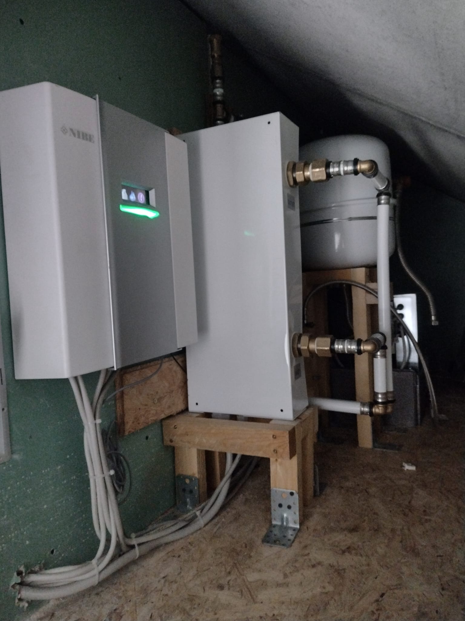 Biała pompa ciepła Nibe zamontowana na drewnianej konstrukcji w pomieszczeniu z zieloną ścianą i widocznymi przewodami. Widoczny zbiornik wyrównawczy.