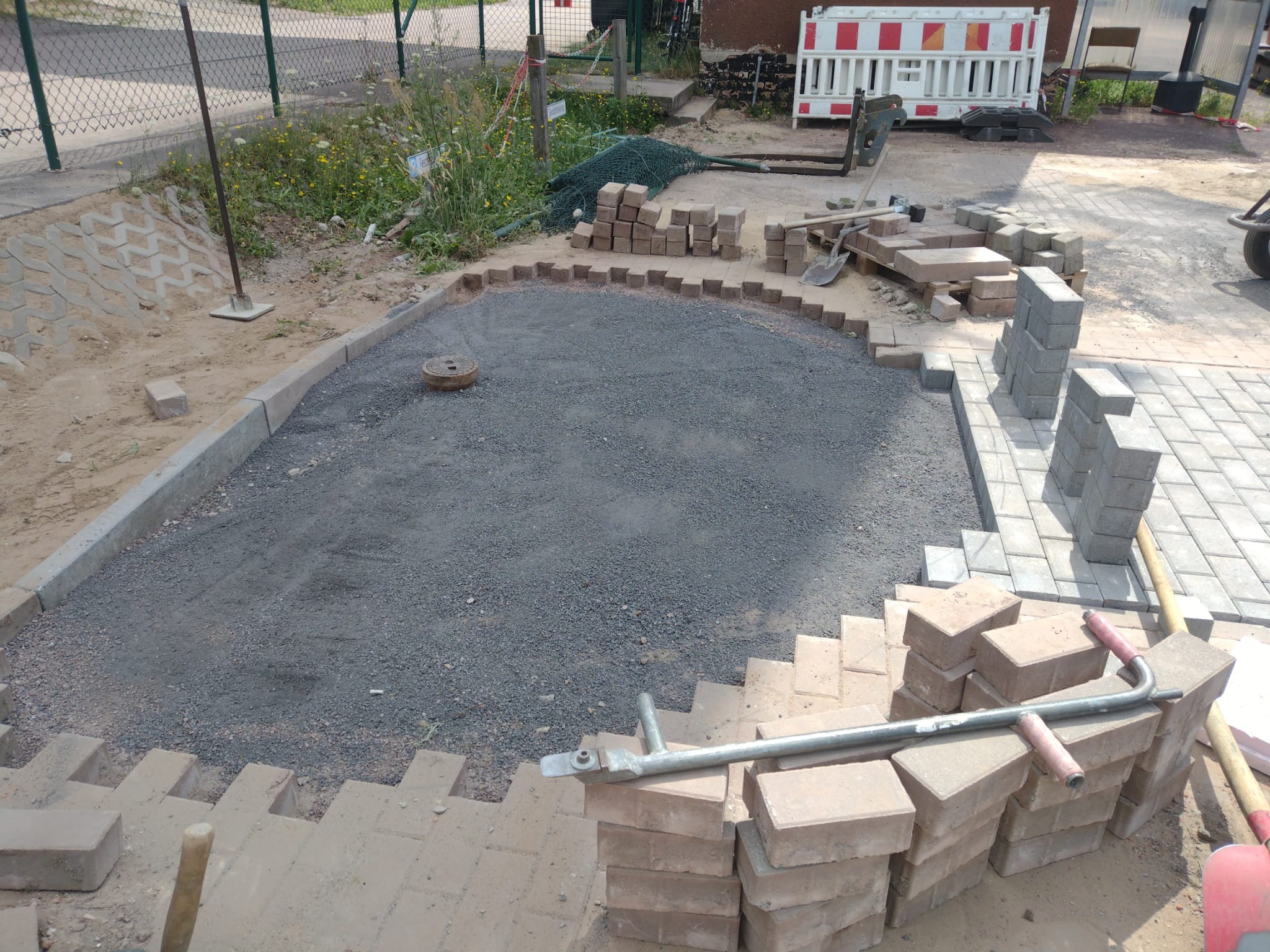 Układanie kostki brukowej: przygotowany teren z podsypką, obrzeża i częściowo ułożona kostka. Widoczne narzędzia i materiały budowlane.