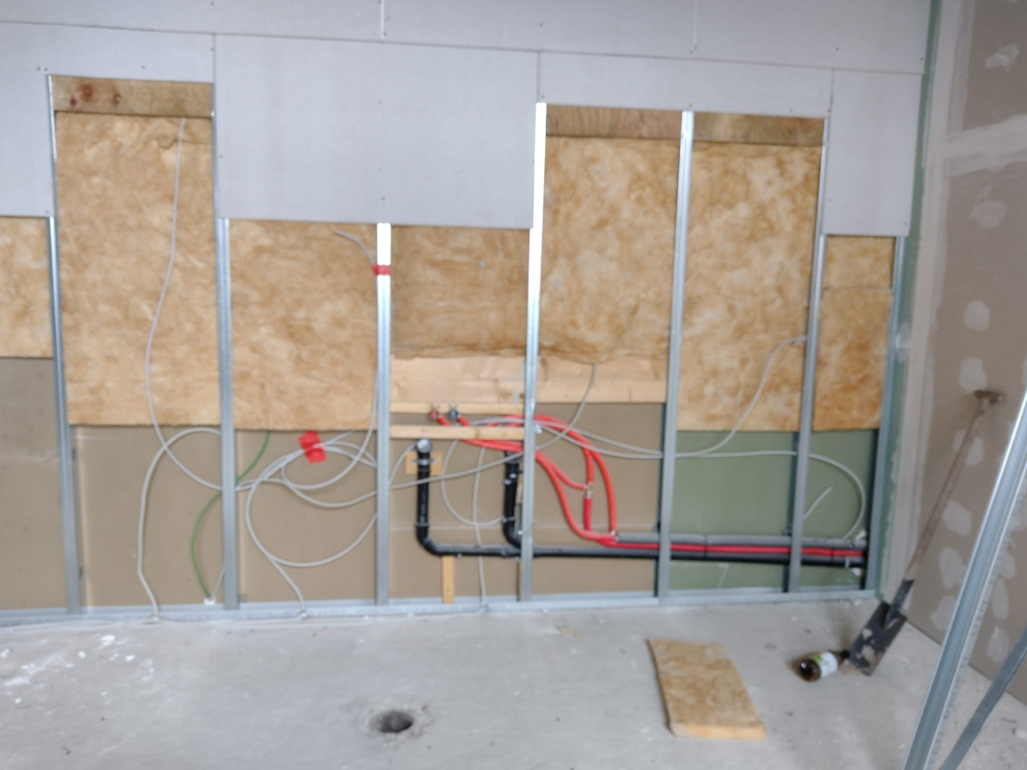 Wnętrze w trakcie prac wykończeniowych: widoczne instalacje hydrauliczne (rury czerwone i czarne) oraz elektryczne (kable białe) w ścianie z izolacją i metalowym stelażem.