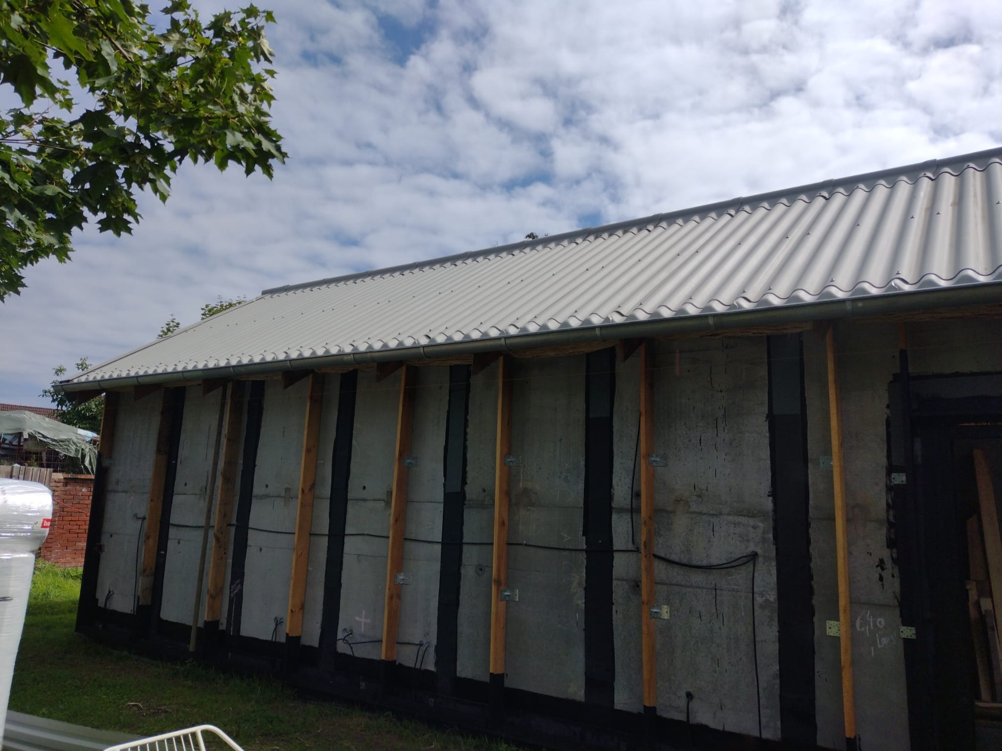 Ściana budynku w trakcie budowy z widocznymi elementami konstrukcyjnymi i izolacją, blaszany dach, fragment zieleni i niebo z chmurami w tle.