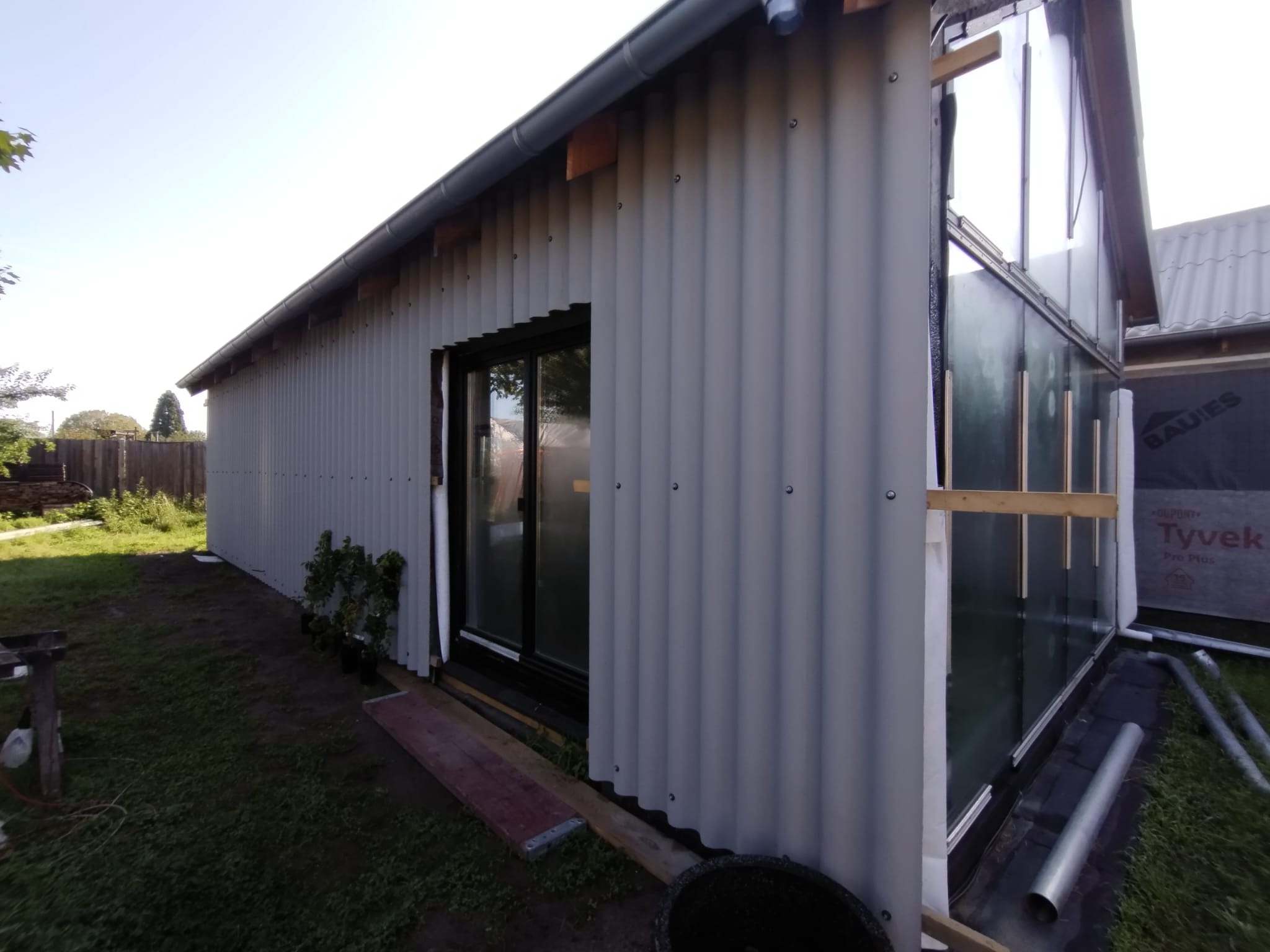 Budowa domu szkieletowego z szarą blachą falistą na elewacji, widoczne okno i folia izolacyjna Tyvek. Ujęcie z zewnątrz, w tle zieleń i fragment dachu.