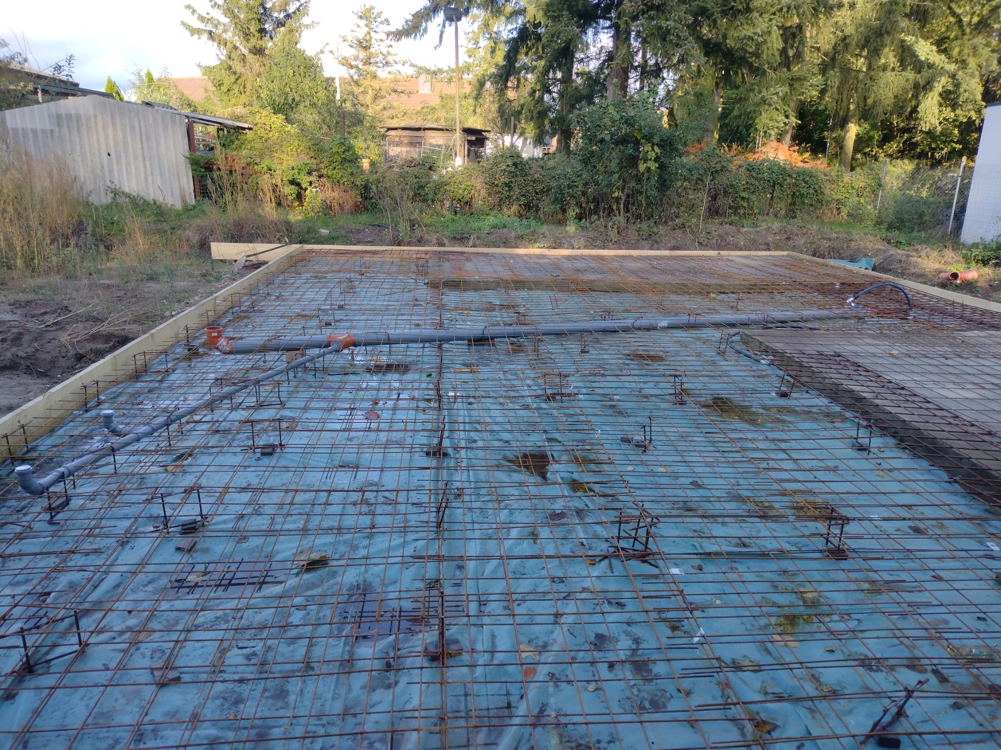 Zbrojenie fundamentu z rurami instalacji podposadzkowej na folii, przygotowane do zalania betonem. Widoczne drewniane szalunki i otoczenie placu budowy.