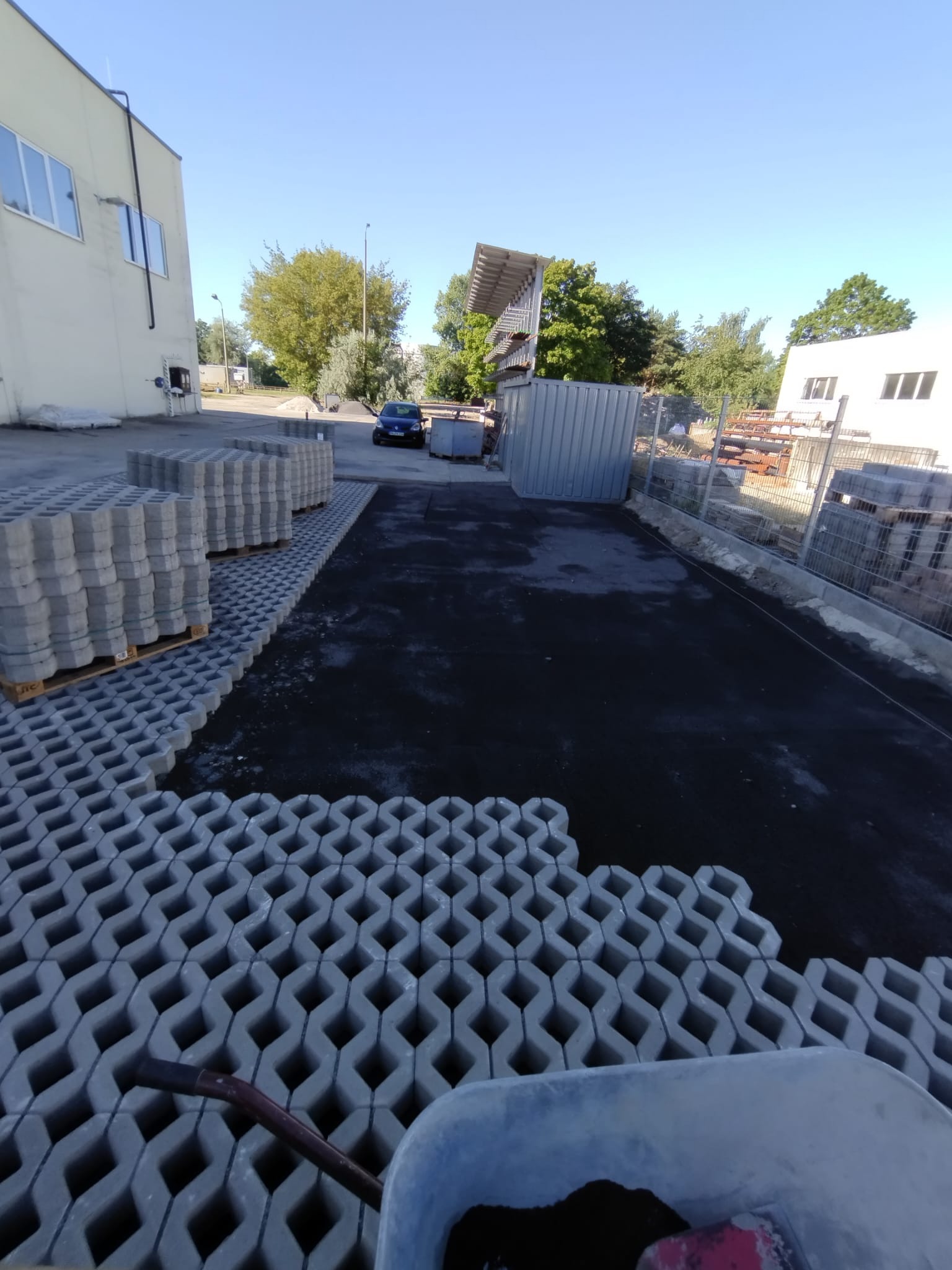 Układanie ażurowych płyt betonowych w Pieszycach. Widoczne palety z kostką i częściowo ułożona nawierzchnia. W tle budynek i drzewa.