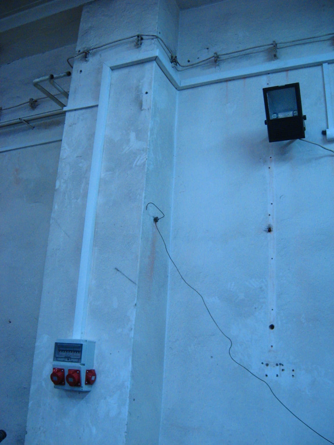 Szara ściana z zamontowaną skrzynką elektryczną z czerwonymi gniazdami, lampą i przewodami. Widoczne elementy instalacji elektrycznej w surowym wnętrzu.
