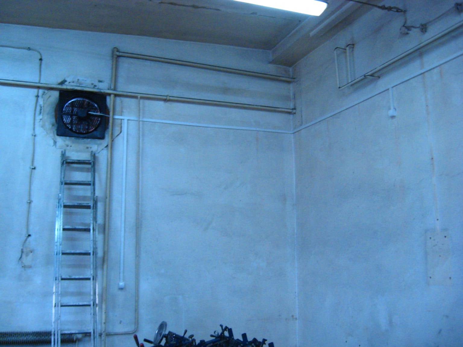 Wnętrze z instalacjami: rury, wentylator z kratką, drabina oparta o ścianę. Widoczne przewody elektryczne i narzędzia na podłodze. Surowe wykończenie ścian.