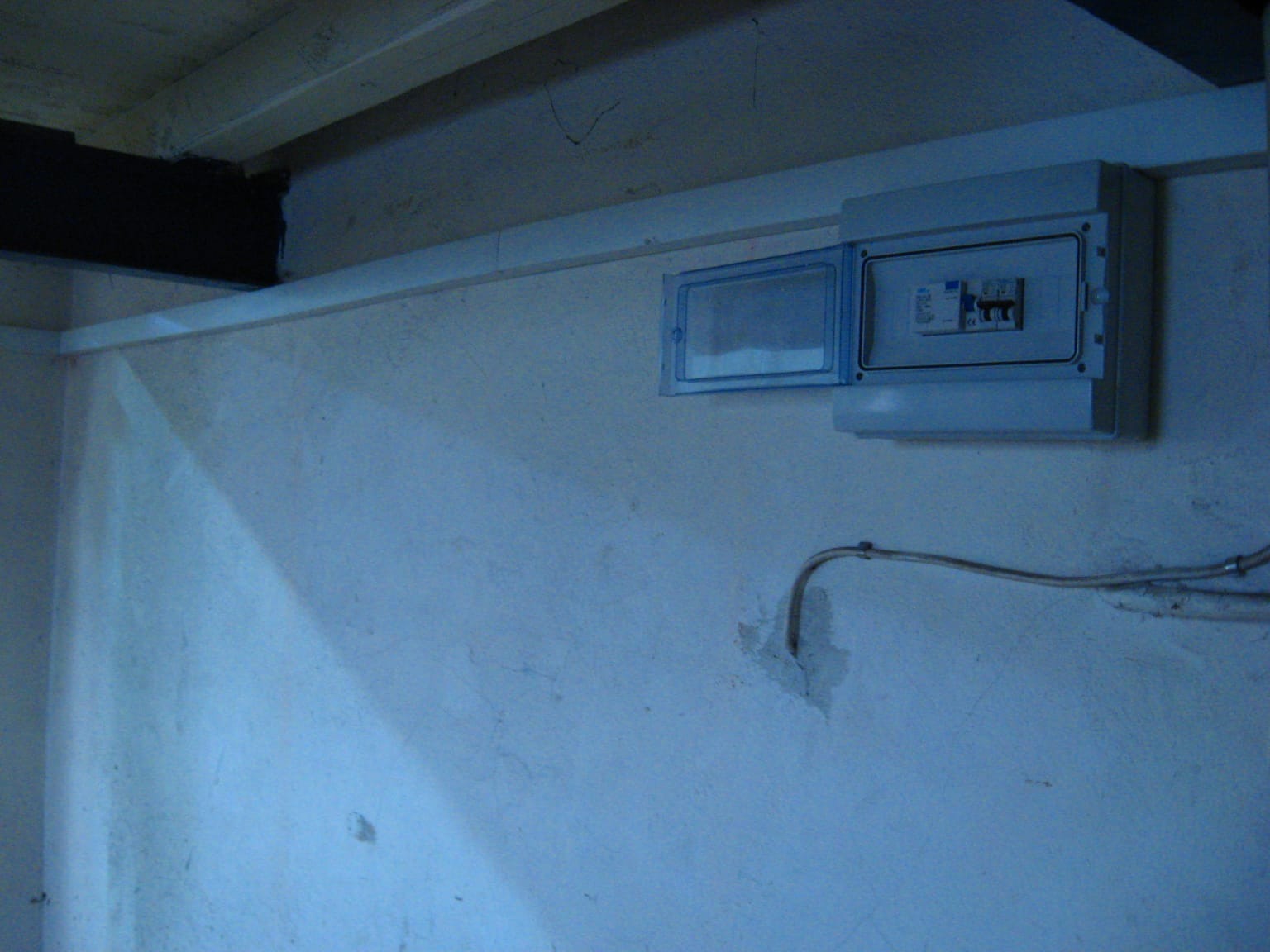 Otwarta skrzynka elektryczna z bezpiecznikami na szarej ścianie wewnątrz budynku. Widoczne kable i fragment sufitu. Ujęcie z dołu.