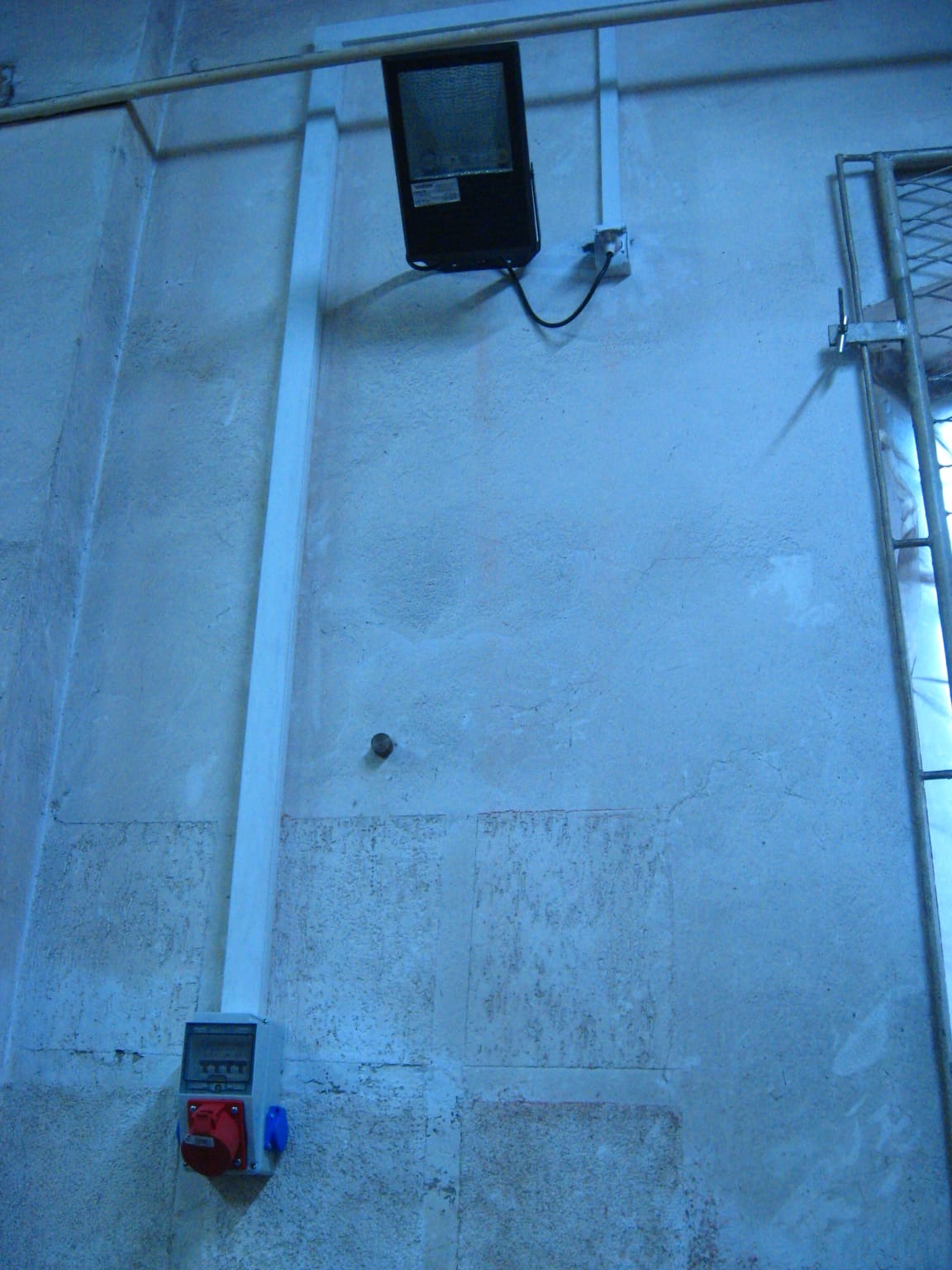 Zamontowana lampa przemysłowa z kablem i skrzynką elektryczną na szorstkiej, pomalowanej ścianie. Widoczna drabina z metalową siatką po prawej stronie.