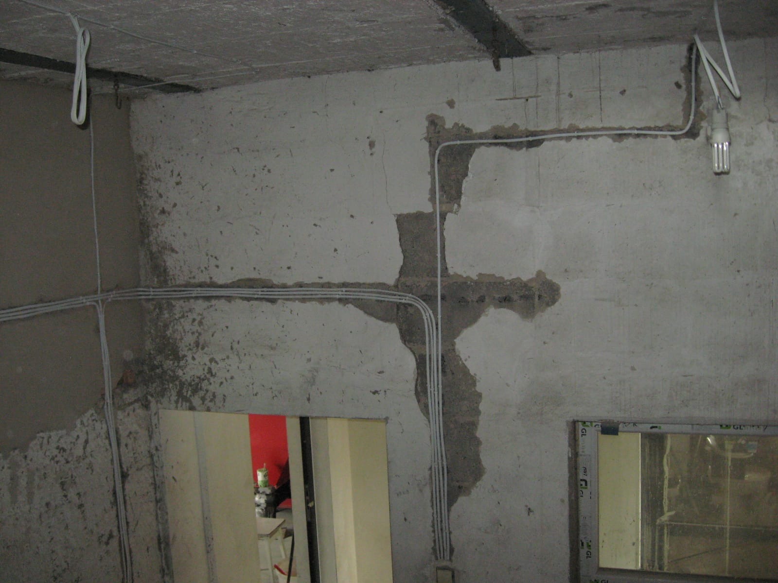 Surowa ściana z widocznymi przewodami elektrycznymi, częściowo ukrytymi w bruzdach. W tle widoczne drzwi i okno. Etap prac instalacyjnych wewnątrz budynku.