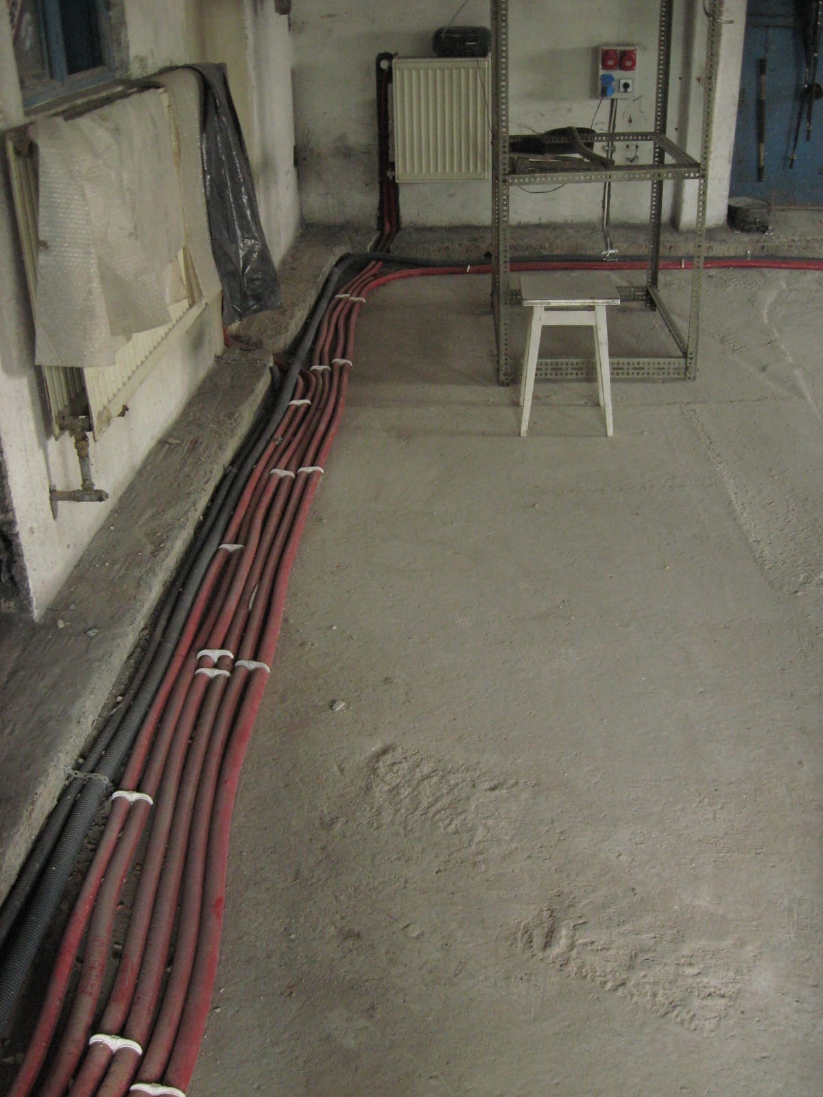 Rury grzewcze i elektryczne ułożone wzdłuż ściany w surowym wnętrzu, widoczne elementy instalacji i metalowy regał. Prace instalacyjne w trakcie realizacji.