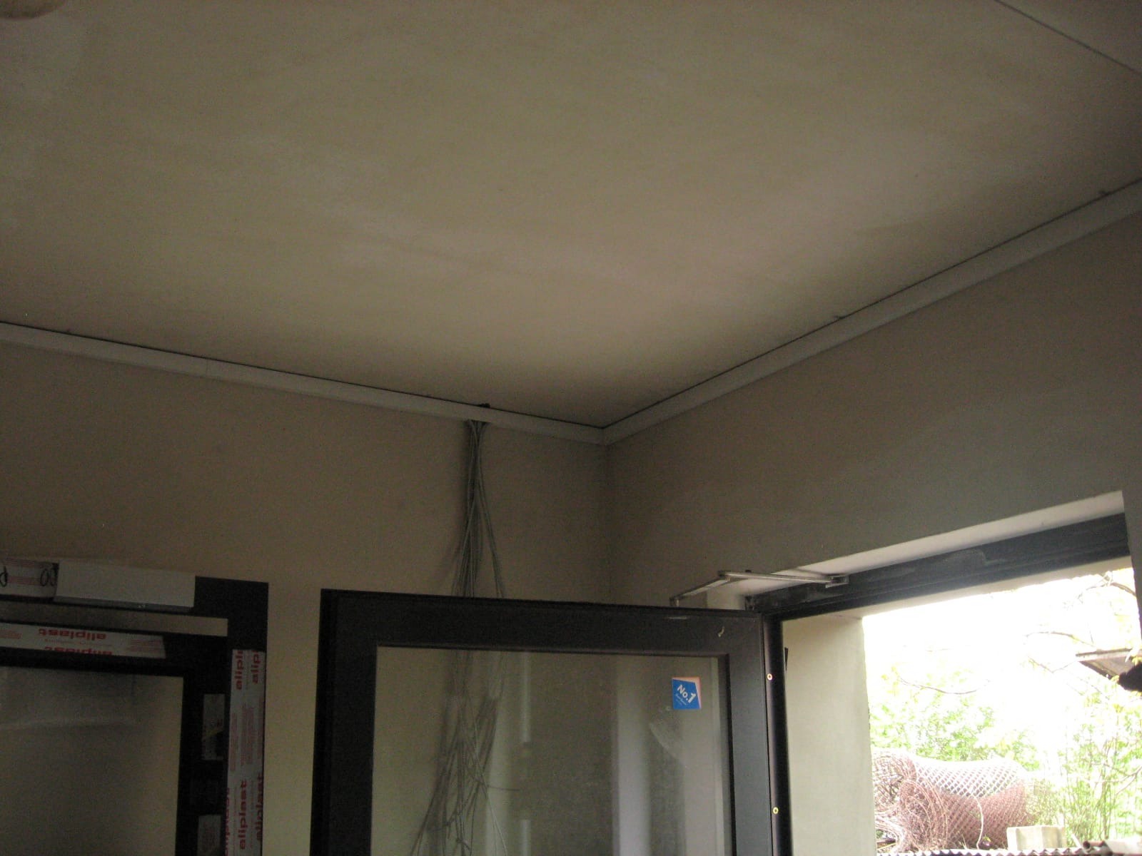 Wnętrze w trakcie wykończenia: widoczne przewody elektryczne zwisające ze ściany, listwa sufitowa i częściowo zamontowane okno z czarną ramą.