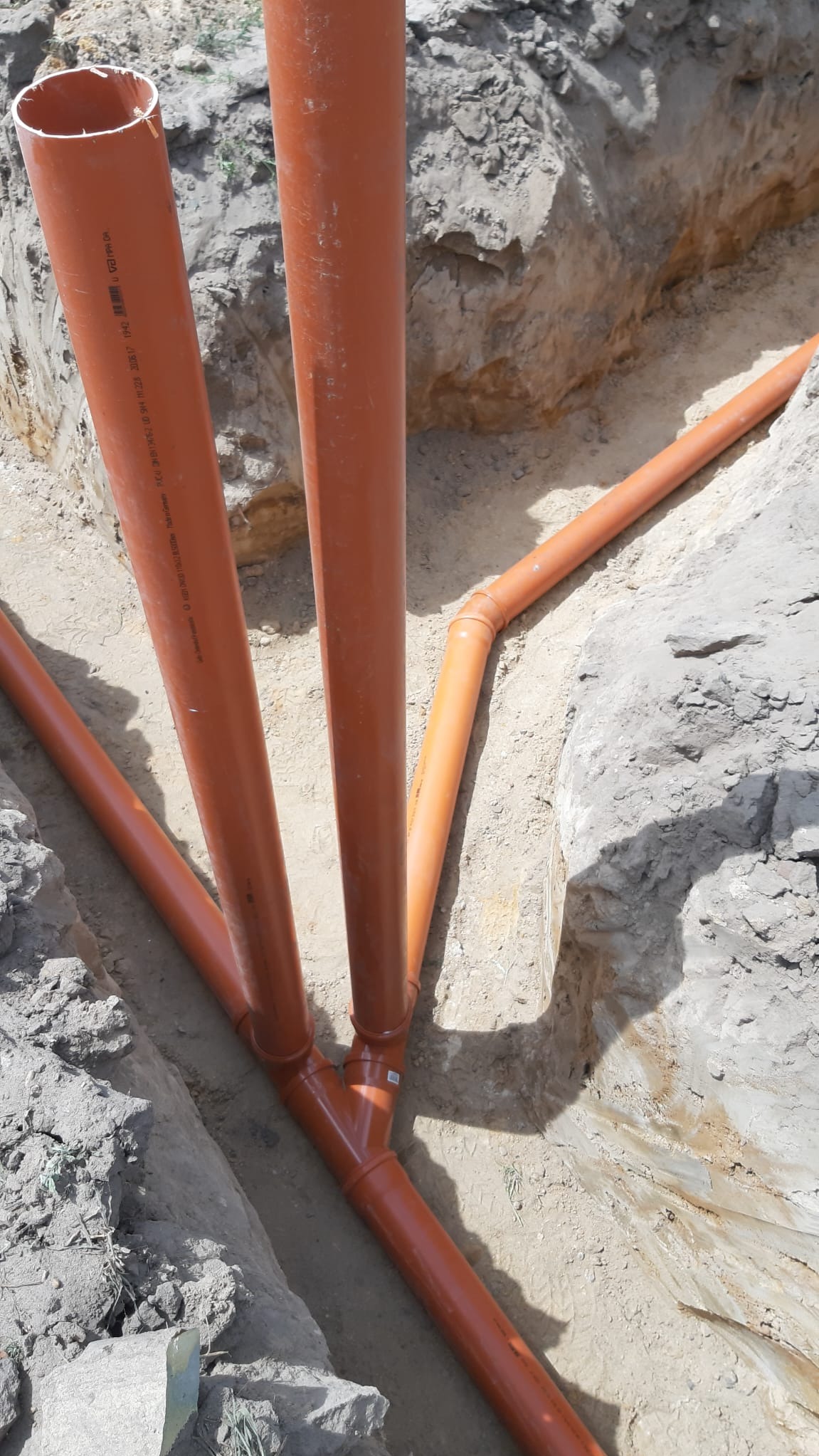 Pomarańczowe rury kanalizacyjne ułożone w wykopie ziemnym, widoczne złącza i fragmenty rur. Instalacja w trakcie montażu, ziemia wokół rur.