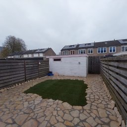 Craft Garden - Ogród z nową trawą i ścieżką z kamieni, otoczony drewnianym ogrodzeniem. W tle biały budynek gospodarczy i zabudowa szeregowa z panelami słonecznymi.