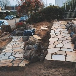 Craft Garden - Ścieżka z nieregularnych płyt kamiennych, obłożona kamieniami, prowadząca przez ogród. W tle widoczne budynki i samochody.