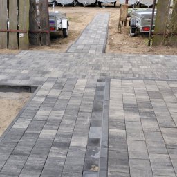 Craft Garden - Szara kostka brukowa ułożona w geometryczny wzór, tworząca ścieżkę. W tle przyczepy i autobus, a po bokach drewniane elementy ogrodzenia.