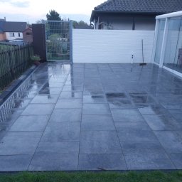 Craft Garden - Nowoczesny taras z szarych, prostokątnych płyt. Widoczne ogrodzenie z metalowej siatki, drewniany płot i biała ściana z cegły. Powierzchnia płyt mokra, odbija światło.