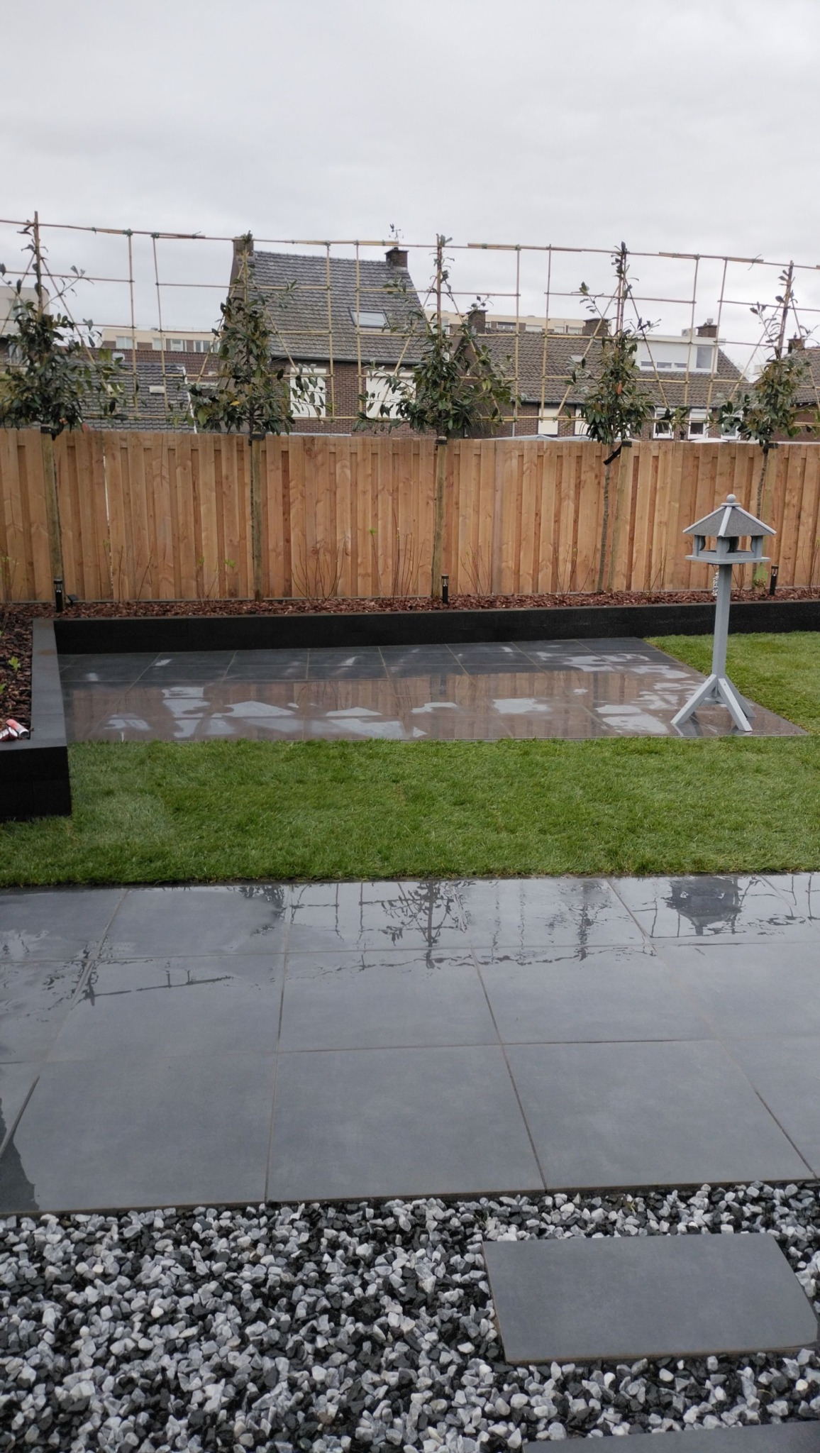 Nowoczesny ogród z szarymi płytami, drewnianym ogrodzeniem i rzędem drzewek. Widoczny karmnik dla ptaków i dekoracyjne kamienie. Całość utrzymana w stonowanej kolorystyce.